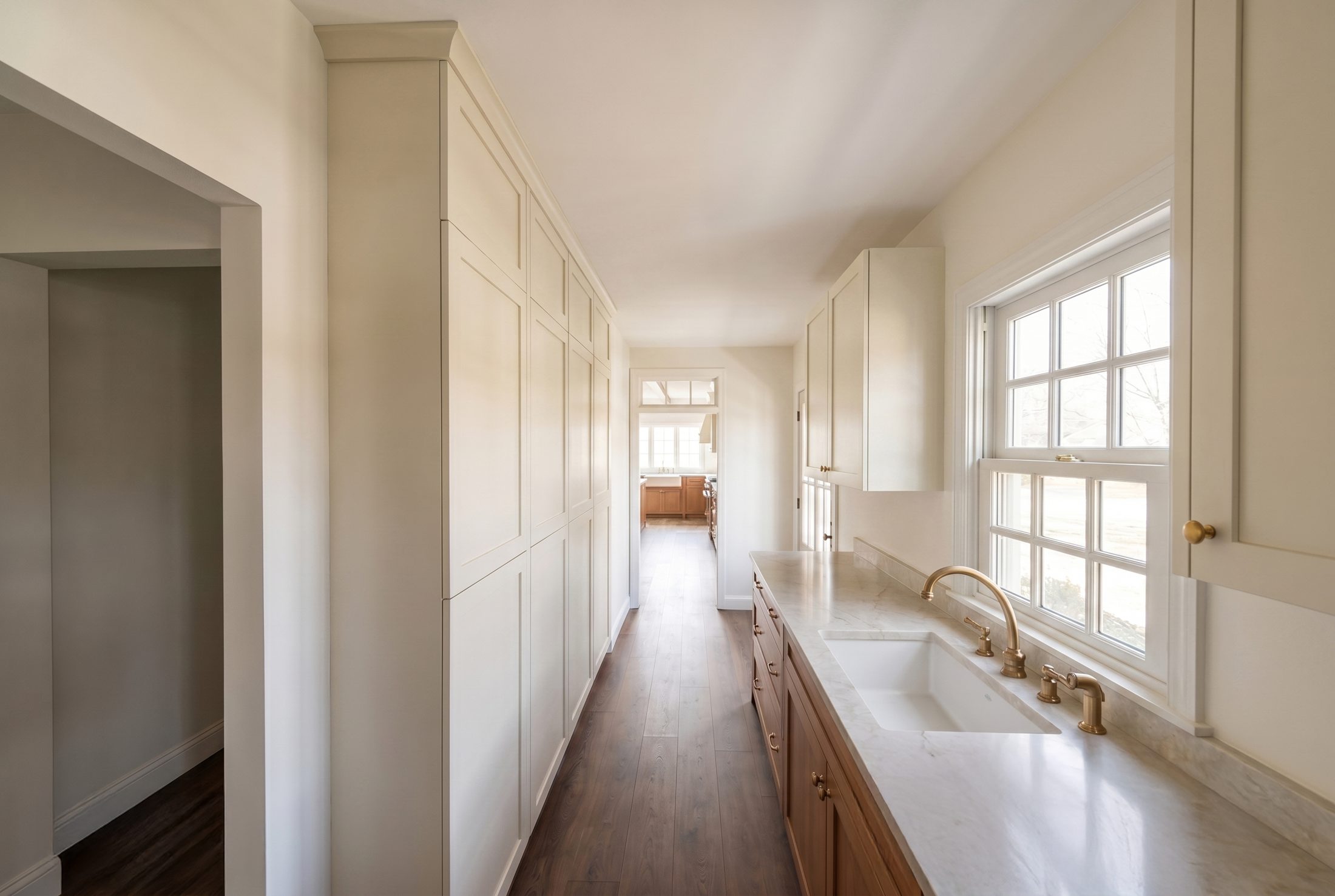 Butler pantry with floor-to-ceiling painted cabinetry, a stone counter, undermount sink and brass fixtures, looking through to the kitchen
