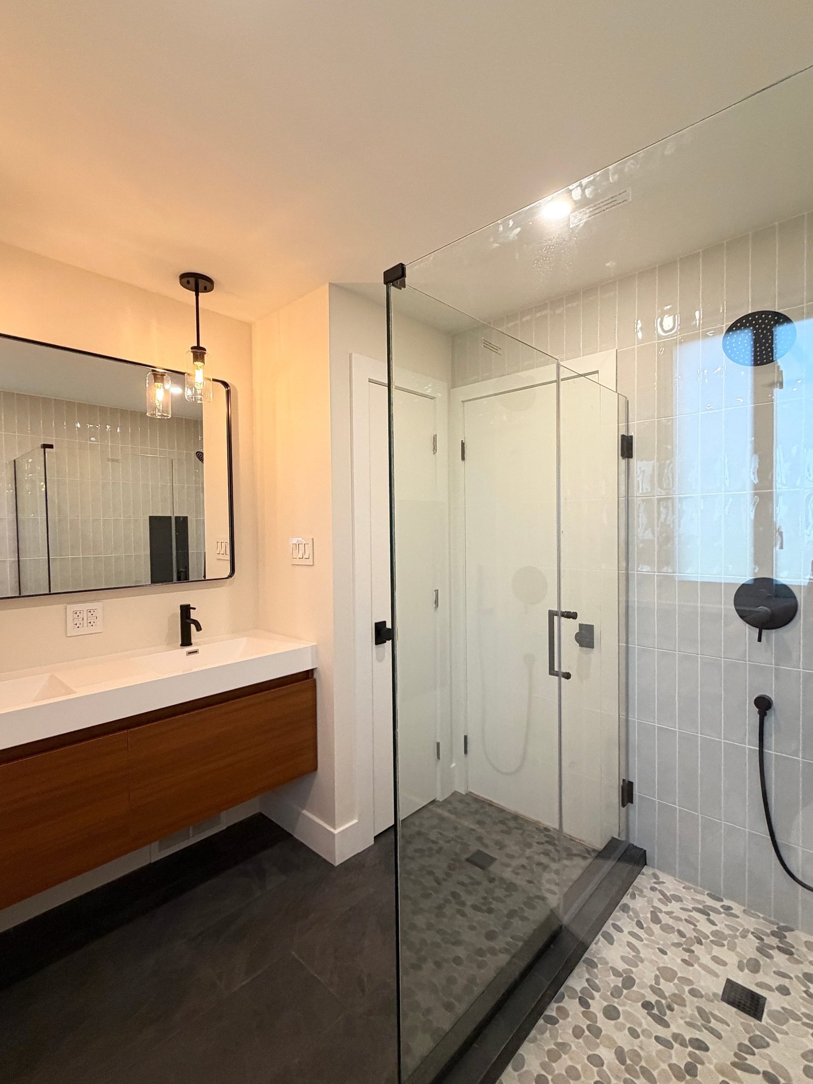 Finished primary bath with a floating wood vanity, black-framed mirror, glass shower and pebble shower floor