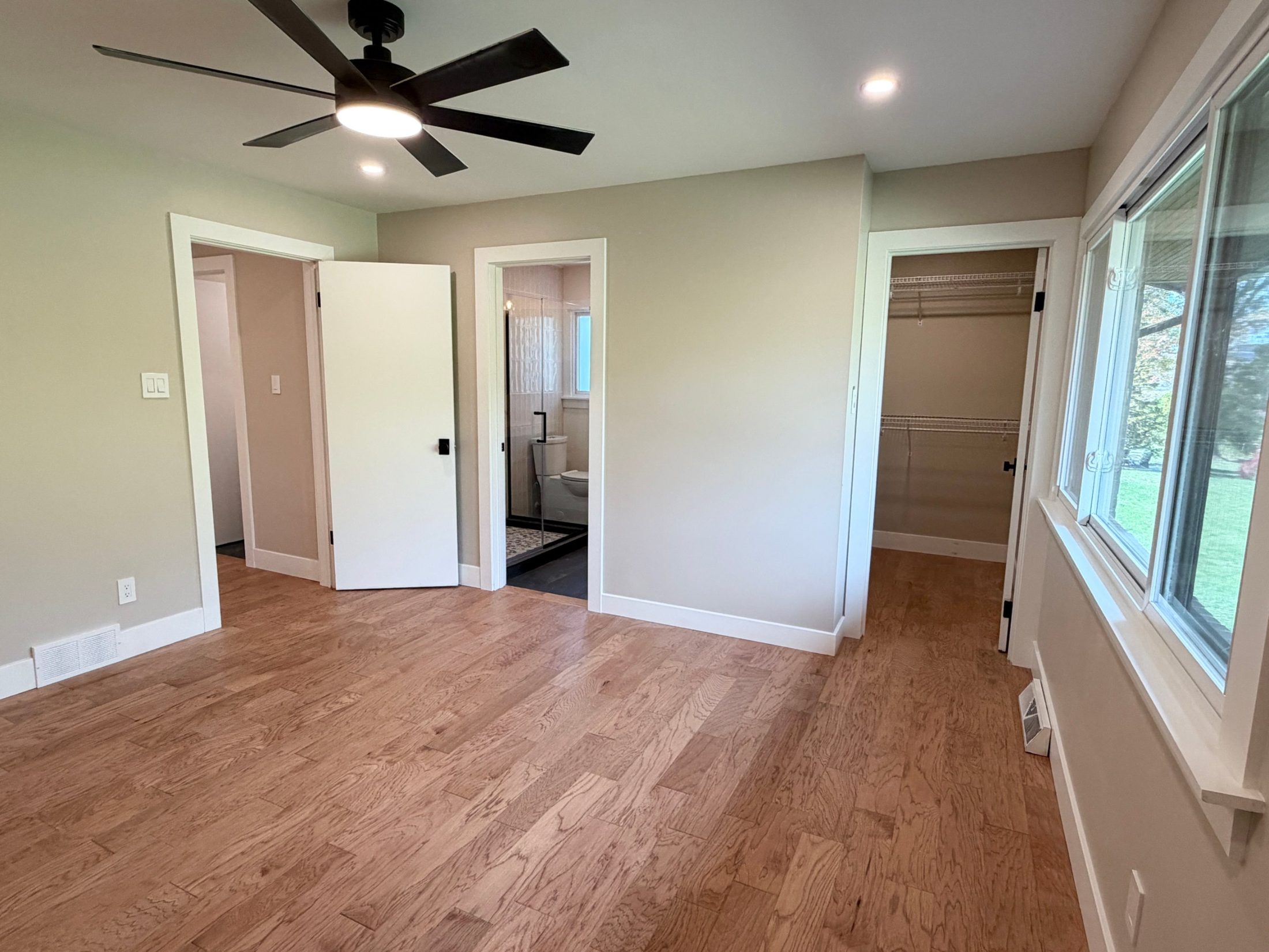 Finished primary bedroom with hardwood floors, a black ceiling fan, a walk-in closet and an en-suite bath