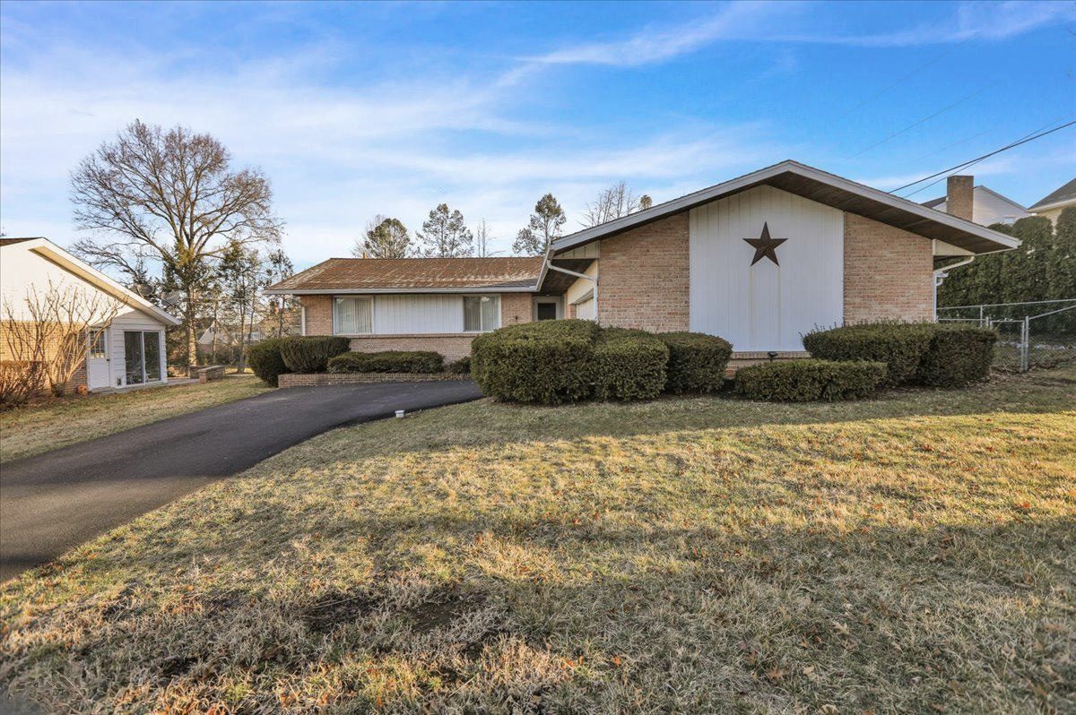The same home before the addition: tan brick ranch with a white board-and-batten gable, a decorative star, shrubs and an asphalt driveway