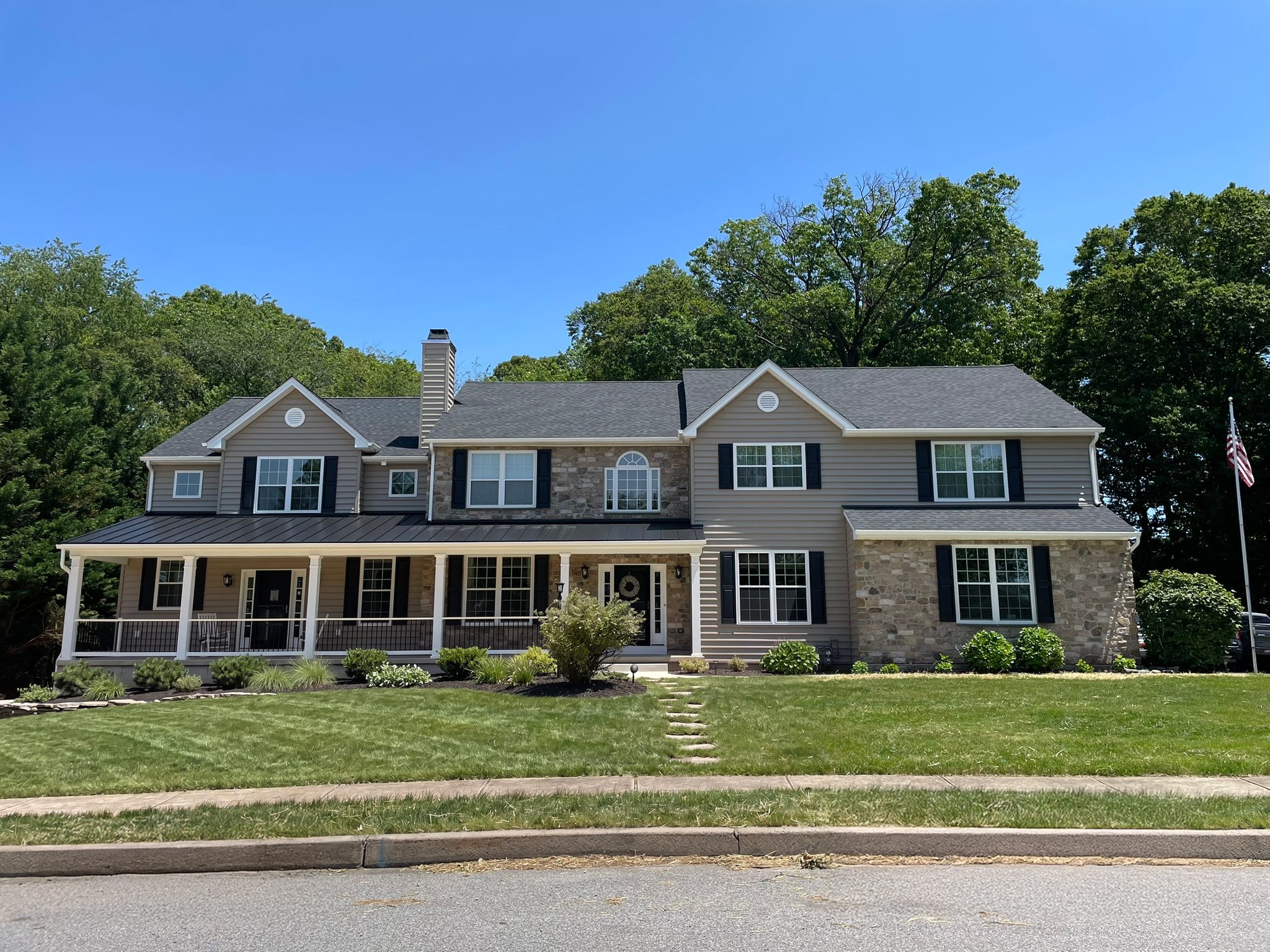 Two-story home after the addition: wraparound front porch with a black standing-seam metal roof, stone and taupe siding, black shutters and a new gabled wing at left