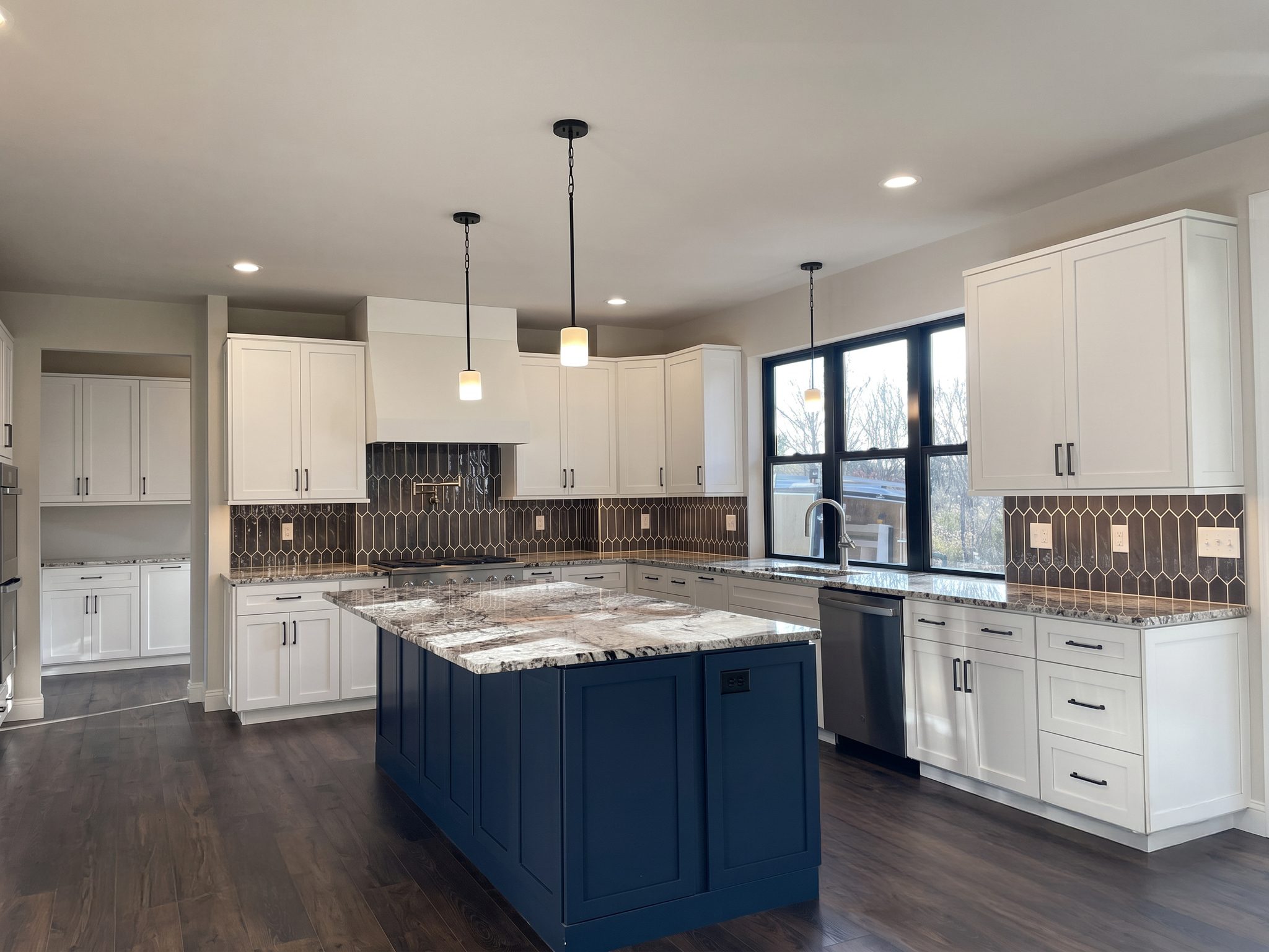 Kitchen with white shaker cabinets, a navy island with granite top, black pendant lights and dark picket-tile backsplash