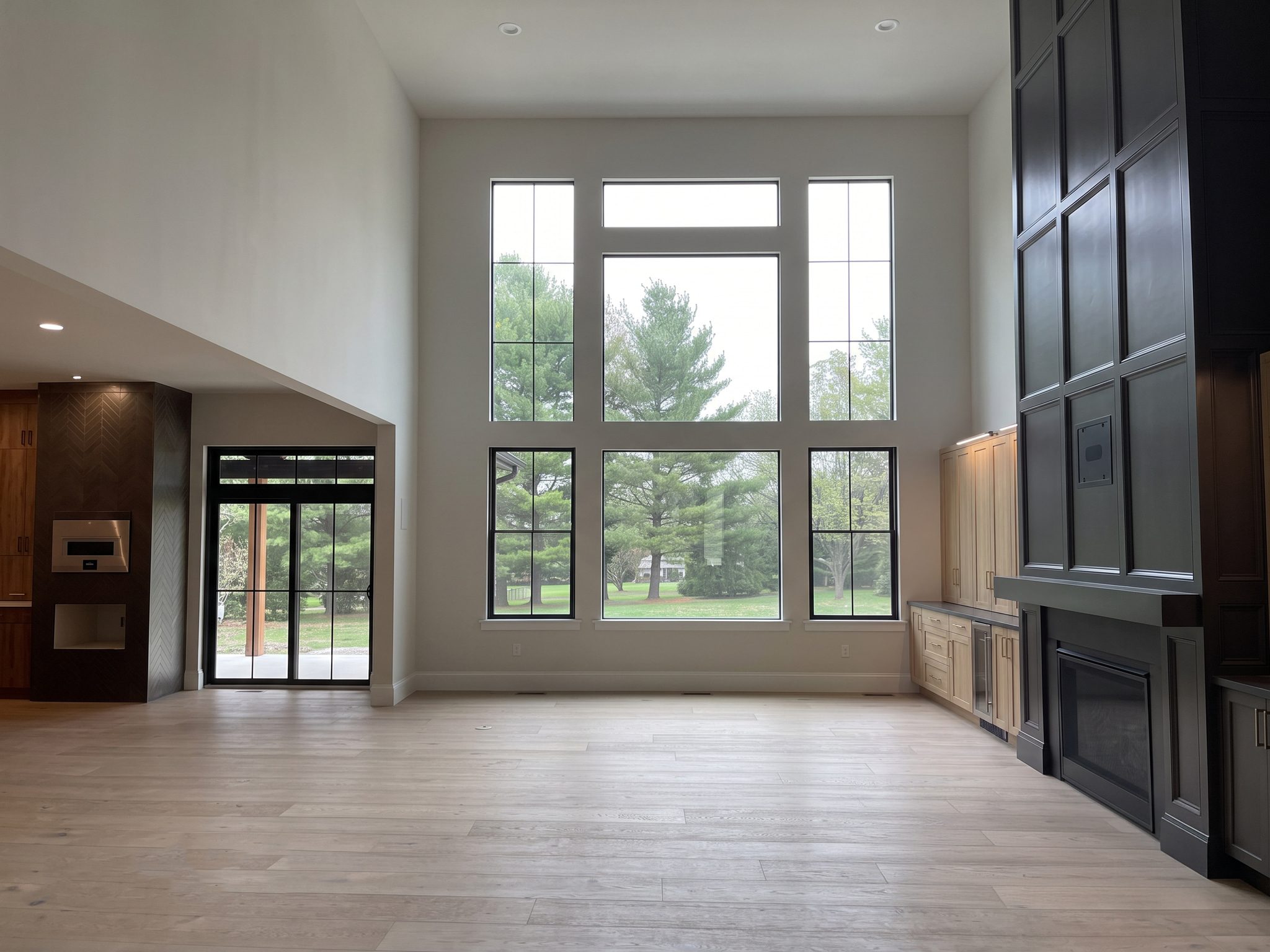 Living room with a tall window wall facing mature pines, wide-plank light oak floors, a tall charcoal paneled fireplace with built-in cabinets, and the kitchen beyond