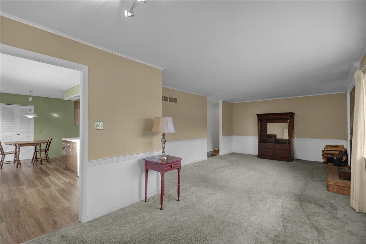 Before: the original living room with tan walls, white wainscoting, gray carpet, a brick hearth and a view into the kitchen