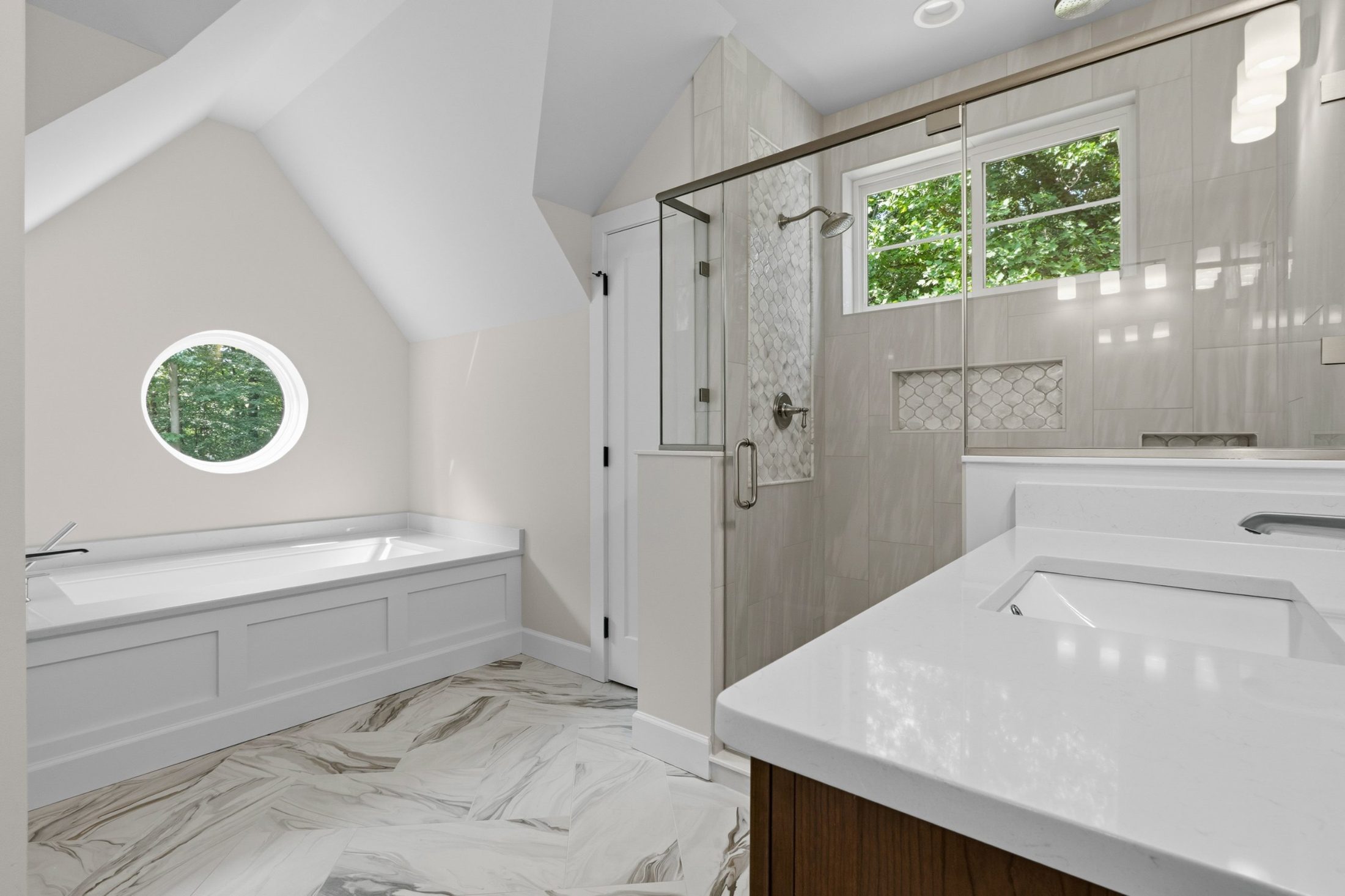 Primary bath with a soaking tub under a round window, glass shower with patterned tile niche and marble-look floor