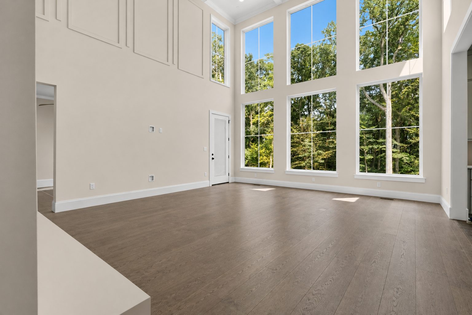 Two-story living room with a wall of tall windows facing the woods, wall paneling and wide-plank wood floors
