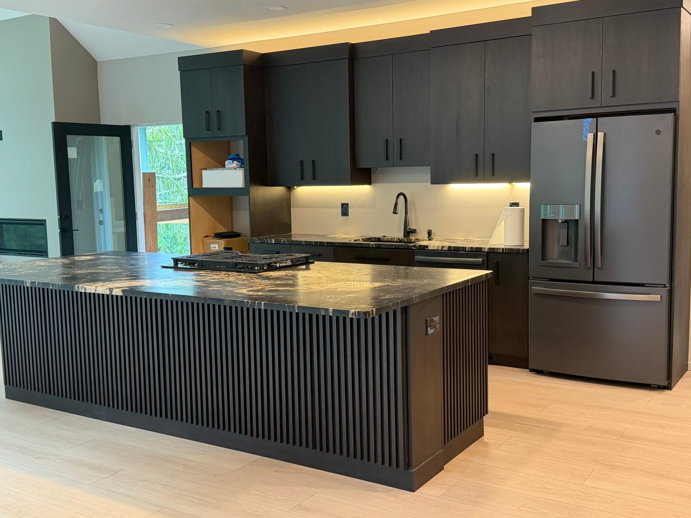 Kitchen with dark slatted island, black stone counters, dark cabinets and cove lighting